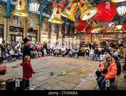 Nelle sale del mercato di Covent Garden, un artista di strada intrattiene la folla tra gli arredi festivi. In un 1 dicembre chiaro ma molto freddo, le luci festive e le decorazioni natalizie illuminano molte aree centrali di Londra affollate di turisti, visitatori e londinesi che si godono la serata. Foto Stock