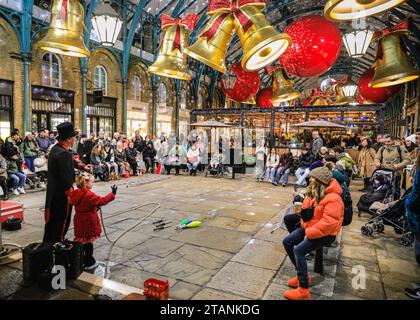 Nelle sale del mercato di Covent Garden, un artista di strada intrattiene la folla tra gli arredi festivi. In un 1 dicembre chiaro ma molto freddo, le luci festive e le decorazioni natalizie illuminano molte aree centrali di Londra affollate di turisti, visitatori e londinesi che si godono la serata. Foto Stock