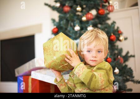Natale, ascolto e un ragazzo che apre un regalo sotto un albero la mattina per festeggiare o per la tradizione. Bambini, regalo e scuotere una scatola con un Foto Stock