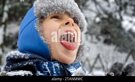 Un ragazzo felice e allegro che cattura fiocchi di neve cadenti con la lingua. Divertimento e gioia durante le vacanze invernali, bambini che giocano all'aria aperta, attività nella neve Foto Stock