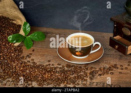 Kaffee Komposition mit Kaffeetasse, Kaffeebohnen in Kaffeesack, historischer Kaffeemühle und Kaffeeblätter auf einem Holztisch Foto Stock