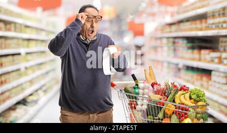 Un uomo maturo scioccato che tiene gli occhiali e guarda un conto all'interno di un supermercato Foto Stock