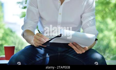 Uomo d'affari che indossa abiti casual seduto in parco e analizza il documento di rapporto delle scorte. Concetto di commercio e documentazione. Foto Stock