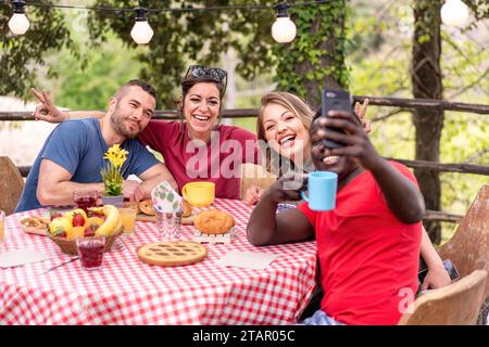 Un gruppo eterogeneo di amici che condividono risate e scattano selfie durante un brunch all'aperto. Foto Stock