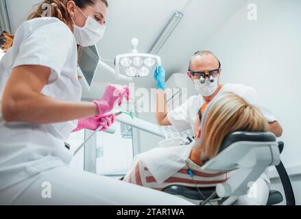 Appuntamento per il paziente in clinica odontoiatrica. Dentista con lenti d'ingrandimento che puntano la luce. Giovane assistente donna pronta per la chirurgia dentaria con strumenti medici Foto Stock