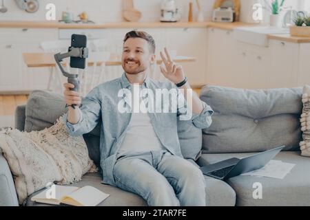 Uomo in abbigliamento casual che registra un video blog, dando un segno di pace con un telefono e una gimbal. Foto Stock