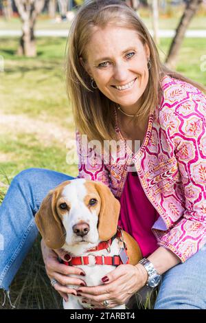 Ritratto di una donna sorridente che guarda la macchina fotografica e abbraccia un cane Beagle seduto sull'erba verde al parco Foto Stock