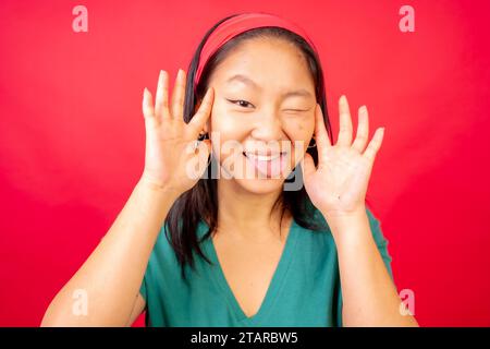 Foto in studio con sfondo rosso di una simpatica giovane donna cinese che tira volti e gesti Foto Stock