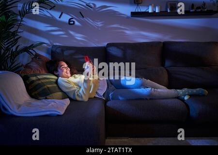 Donna latina sorridente che guarda il cellulare mentre si rilassa in salotto a casa. Giovane donna che usa il cellulare Foto Stock