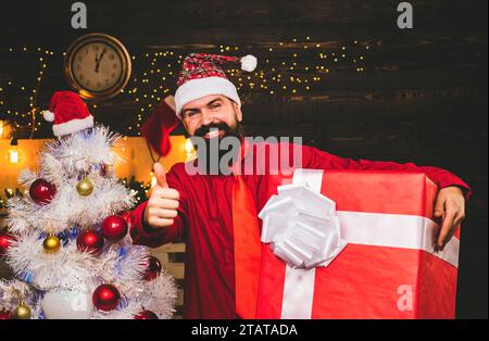 L'uomo di Natale in abito rosso alla moda regge un regalo. Regalo creativo. Hipster Babbo Natale. Babbo natale di Natale con regalo. Foto Stock