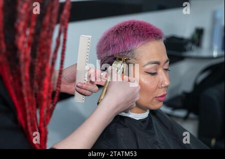 Il parrucchiere rasa il tempio di una cliente femminile. Donna asiatica con capelli corti rosa in barbiere. Foto Stock