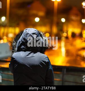 Donna che indossa un cappotto invernale mentre guarda le luci della città in un giorno piovoso di notte. Foto Stock