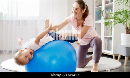 Un bambino sorridente che si diverte sdraiandosi sul fitball e facendo stretching con la madre. Concetto di assistenza sanitaria infantile, bambini sport e yoga Foto Stock