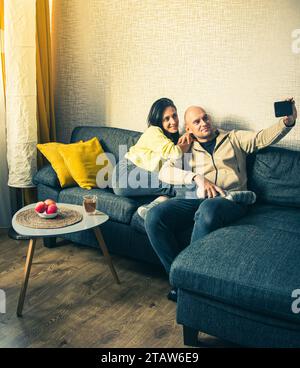 Coppia felice innamorata che si fa un autoritratto con un cellulare. Il divano di casa ti consente di rilassarti dopo il lavoro nel soggiorno Foto Stock