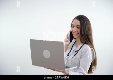 giovane medico donna parlare al telefono e guardare conferenza online in portatile tempo di quarantena trattamento di comunicazione correzione trattamento sul falendoscopio collo su studio di sfondo bianco Foto Stock