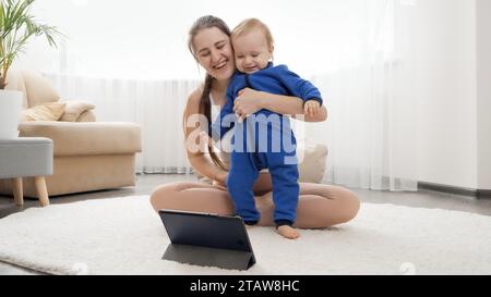 Foto divertente di un bambino che guarda video sul tablet mentre la madre fa esercizi di fitness con lui. Assistenza sanitaria familiare, stile di vita attivo, pa Foto Stock
