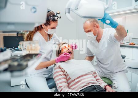 Appuntamento per il paziente in clinica odontoiatrica. Dentista donna in lenti d'ingrandimento facendo prevenzione dei denti utilizzando strumenti medici. Giovane assistente che punta la luce. Foto Stock
