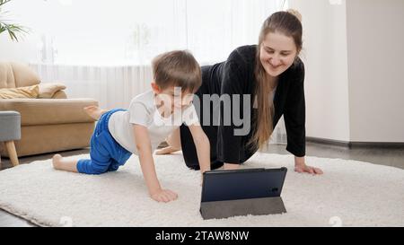 Sorridente ragazzo con madre che guarda lezioni di fitness online su tablet e fa esercizi sportivi a casa. Assistenza sanitaria familiare, stile di vita attivo, genitorialità Foto Stock