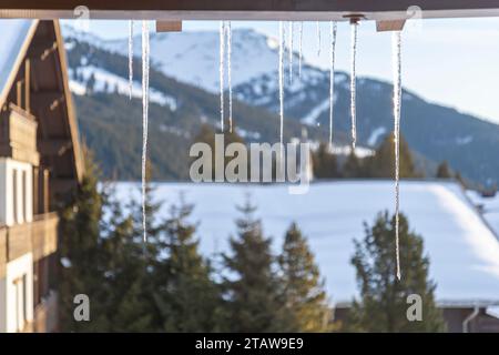 Ghiaccioli appesi dal tetto su uno sfondo sfocato di montagne e i tetti delle case sono ricoperti di neve Foto Stock