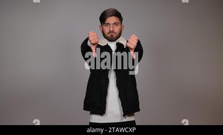 Un giovane che posa con il pollice verso il basso ed esprime antipatia su sfondo grigio. Foto Stock