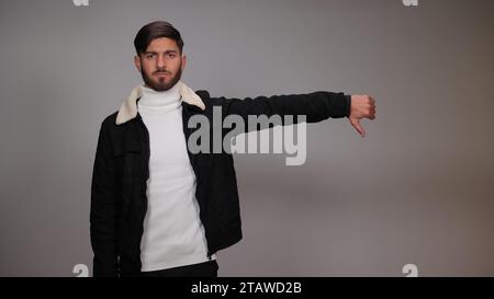 Un giovane che posa con il pollice verso il basso ed esprime antipatia su sfondo grigio. Foto Stock