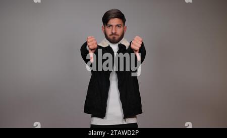 Un giovane che posa con il pollice verso il basso ed esprime antipatia su sfondo grigio. Foto Stock
