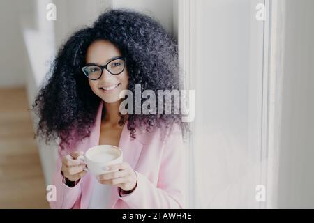 Donna sorridente con capelli ricci che si gode una tazza di caffè con un blazer rosa. Foto Stock