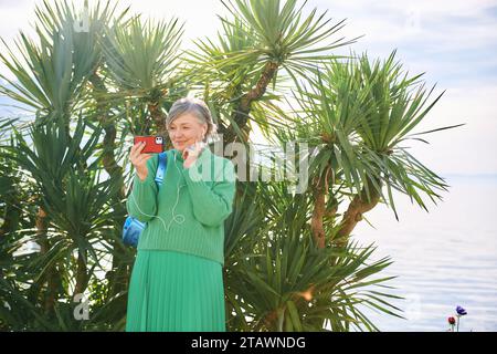 Ritratto di una donna di mezza età che si gode una bella giornata all'aperto, si rilassa sulla panchina di fronte al lago di montagna, tiene il telefono, effettua videochiamate Foto Stock