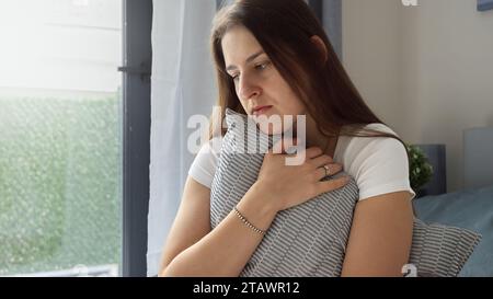 Con le lacrime agli occhi, la giovane donna siede sul letto, le braccia avvolte intorno a un cuscino in un abbraccio stretto. Il concetto di angoscia emotiva, solitario Foto Stock