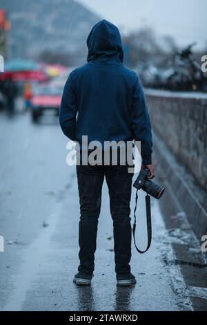 Un fotografo che indossa una felpa con cappuccio, regge una macchina fotografica e si trova in strada. Foto Stock