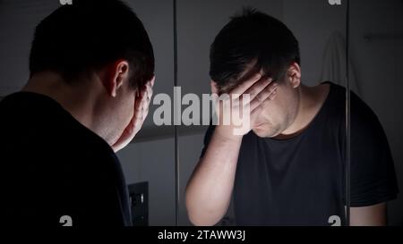 Un uomo triste e frustrato che guarda allo specchio e sospira. Concetto di depressione, suicidio, stress, malattia mentale, solitudine e frustrazione. Foto Stock