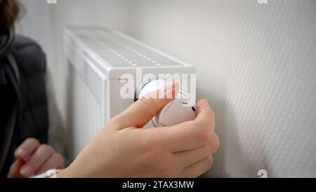 Primo piano di una donna che regola il termostato del radiatore di riscaldamento e riscalda le mani. Concetto di crisi energetica, bollette elevate, sistema di riscaldamento rotto, economia e. Foto Stock