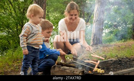 Due ragazzi che guardano la madre che cucina le salsicce sul fuoco nella foresta. Attività ricreative, bambini in campeggio, vacanze in famiglia nella natura Foto Stock