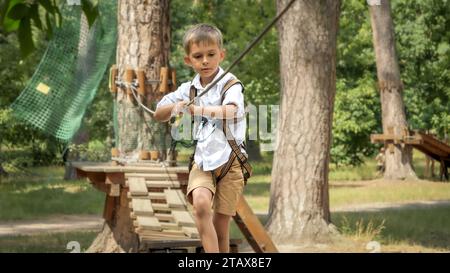 Giovane ragazzo che attraversa un ponte di legno traballante nel parco avventura nella foresta. Infanzia attiva, stile di vita sano, bambini che giocano all'aperto, bambini in natura Foto Stock