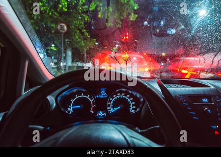 Vista interna di un'auto, dal punto di vista del conducente, si è fermata al semaforo in una giornata di pioggia. Luci plancia, parabrezza a goccia di pioggia. Foto Stock