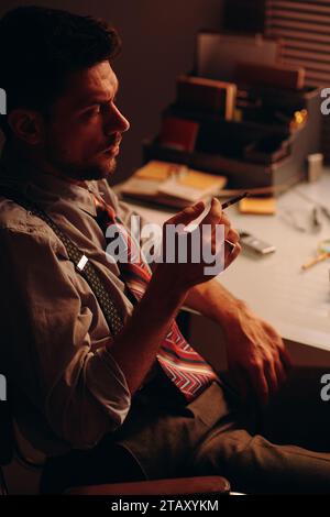 Vista laterale di un giovane uomo d'affari stanco con sigaretta fumante in mano seduto davanti al posto di lavoro di fronte alla macchina fotografica e in pausa Foto Stock