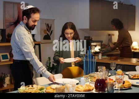 Un uomo maturo e sua figlia mettono piatti e altre posate a tavola con piatti tradizionali ebraici fatti in casa preparati per Hanukkah Foto Stock