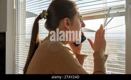 Giovane donna preoccupata che parla al telefono e guarda attraverso le tende. Testimone del crimine, spiare dalla finestra, sbirciare per strada Foto Stock