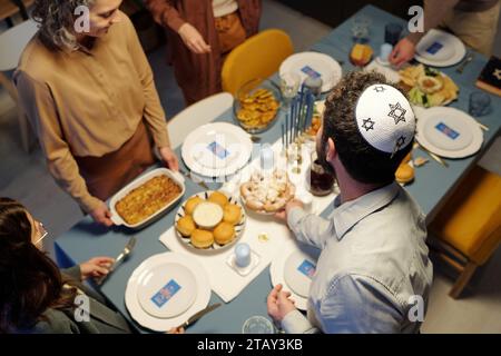 Alto angolo di uomo ebreo che indossa kippah bianco con la stella di David ricamata che mette piatto con spuntino fatto in casa sul tavolo servito Foto Stock