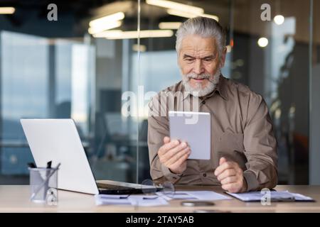 Uomo d'affari di sesso maschile con i capelli grigi lavora in ufficio, siede a un tavolo, tiene un tablet in mano e parla durante una videochiamata con partner e clienti. Foto Stock