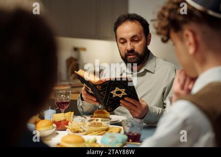 Concentrati sul rabbino barbuto e sul capo della famiglia ebraica che legge testo dalla Torah mentre siede al tavolo servito di fronte ai suoi due figli Foto Stock