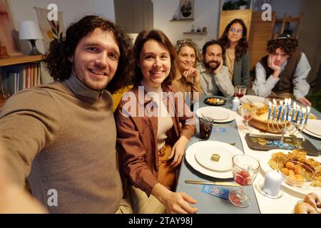 Felice giovane coppia che scatta selfie con i membri della famiglia seduti a un tavolo festivo servito e gustando una cena Hanukkah Foto Stock