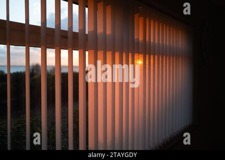 Poco profonda focalizzazione sulle tende verticali delle finestre con un bagliore dorato al tramonto. C'e' una vista del mare e del bagliore del sole che tramonta. Foto Stock