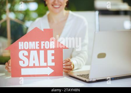 Affari immobiliari ecologici. Primo piano sull'agente immobiliare donna di mezza età moderna felice in moderno ufficio verde in camicetta bianca con casa in vendita segno a Foto Stock