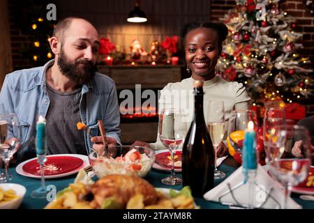 Donna afro-americana a cena festiva le vacanze di dicembre con gli amici che si radunano intorno al tavolo e mangiano cibo tradizionale. Gruppo di persone felici che si divertono la vigilia di natale a casa. Foto Stock