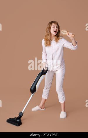 Ritratto di una bella giovane donna in pigiama che canta con la spazzola per capelli mentre usi l'aspirapolvere su sfondo beige Foto Stock