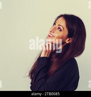 Bella donna d'affari sorridente che guarda in alto con la mano sotto il viso in camicia in cotone nero su sfondo blu chiaro con e. Foto Stock