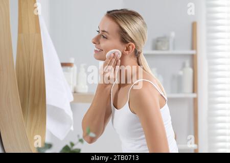 Donna sorridente che si toglie il trucco con un batuffolo di cotone davanti allo specchio in bagno Foto Stock