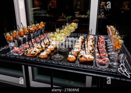 Una varietà di spuntini salati e dolci su un piatto nero e argento, come gamberi, salmone, formaggio, cracker, e frutta, servita con bicchieri da vino. Foto Stock