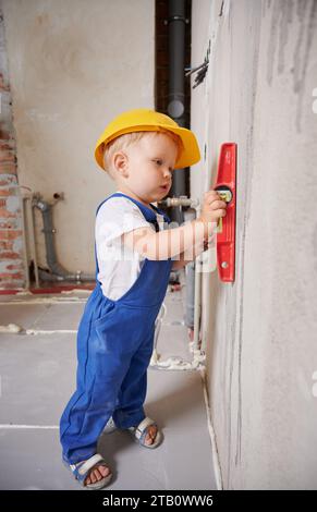 Lavoratore edile per bambini che controlla la superficie della parete con una livella a bolla d'aria. Tutta la lunghezza del bambino in tute da lavoro che utilizza uno strumento livellato durante la preparazione della parete per i lavori di riparazione a casa durante la ristrutturazione. Foto Stock